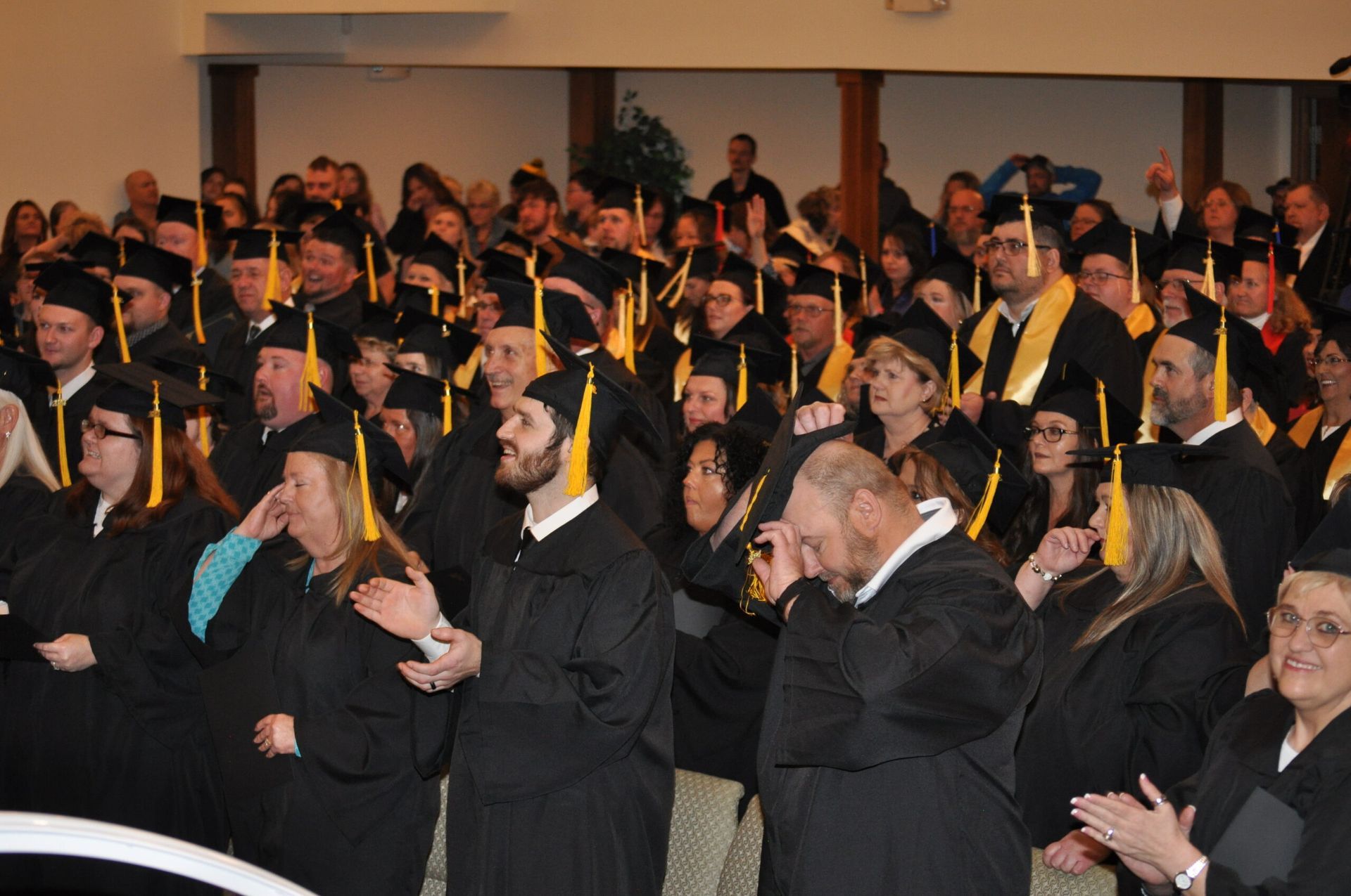 Graduation ceremony: Crowd in black caps and gowns, some with gold stoles, indoors with beige walls.