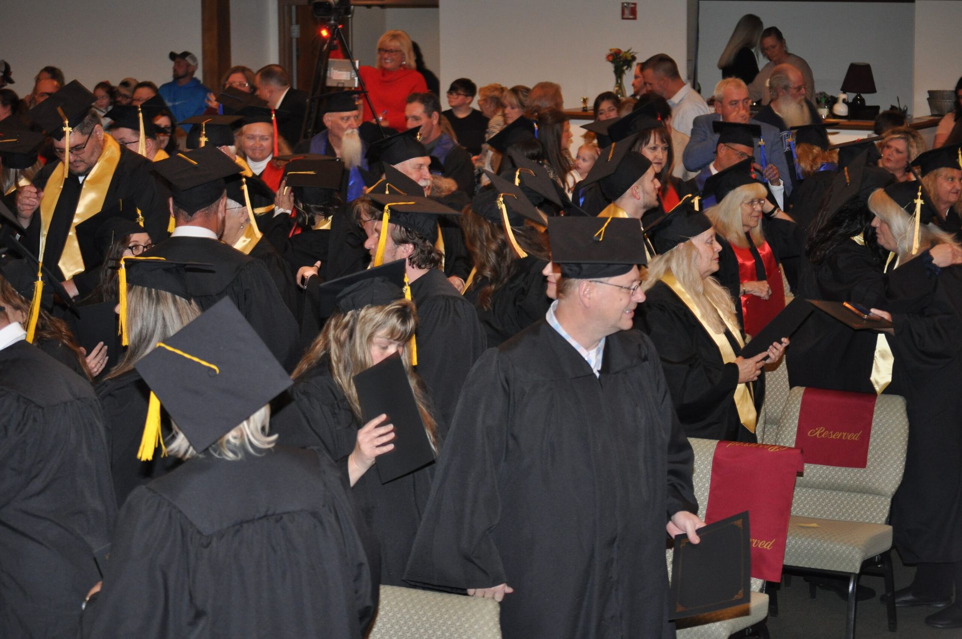 Graduates in black caps and gowns at a ceremony, many holding diplomas; audience in background.