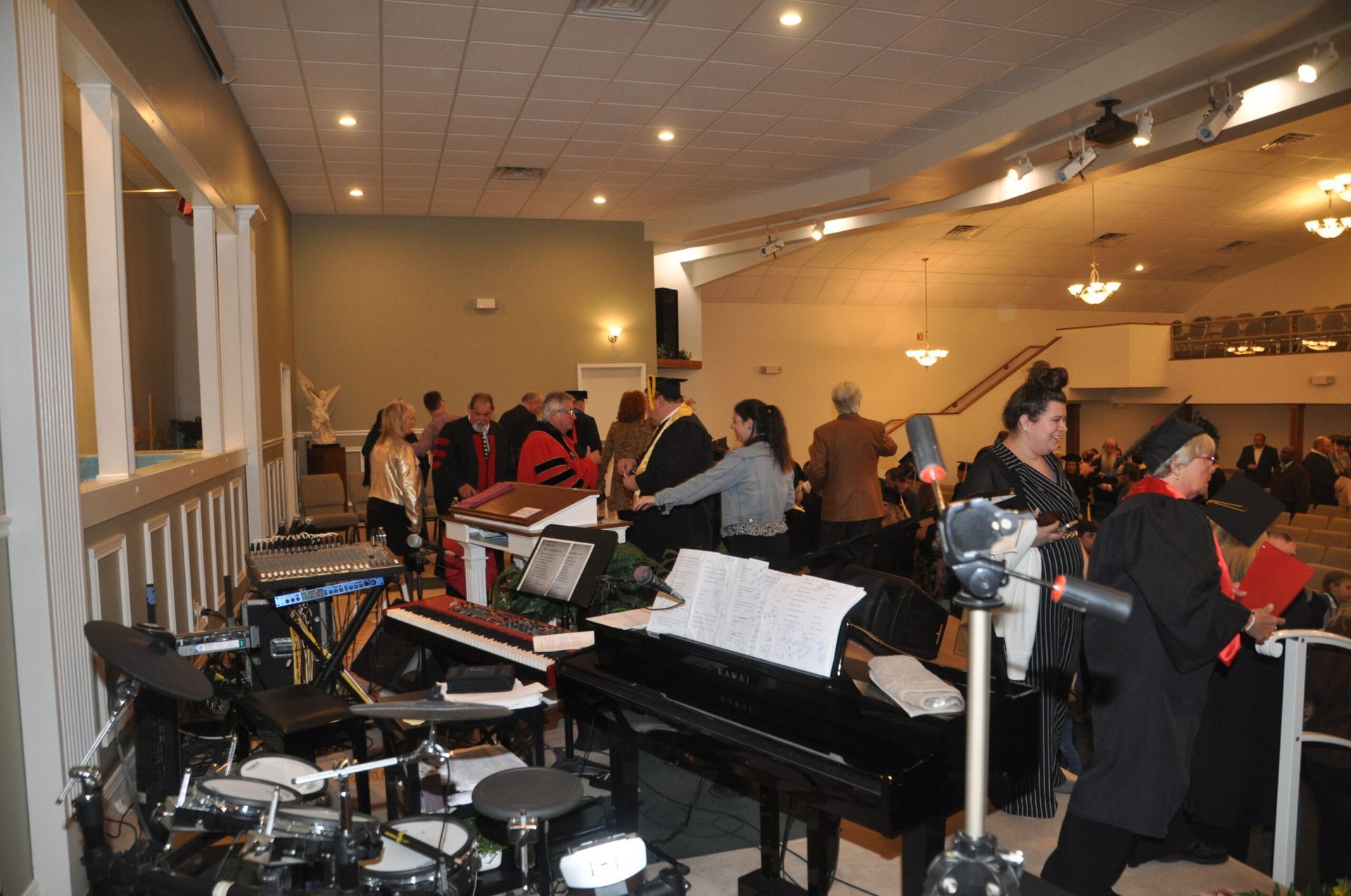 Band setup and people gathered in a large hall with a balcony; lights and musical instruments visible.