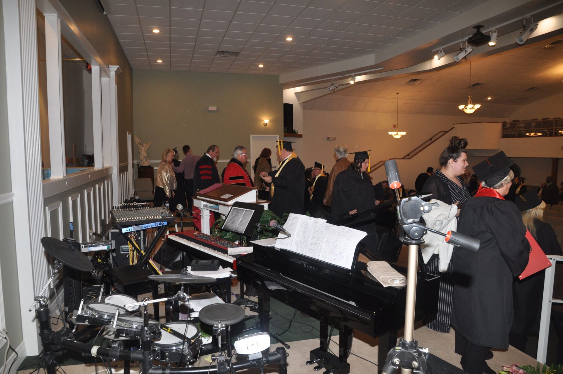 Band setup in a venue, with piano, drums, keyboards, and people in formal attire.