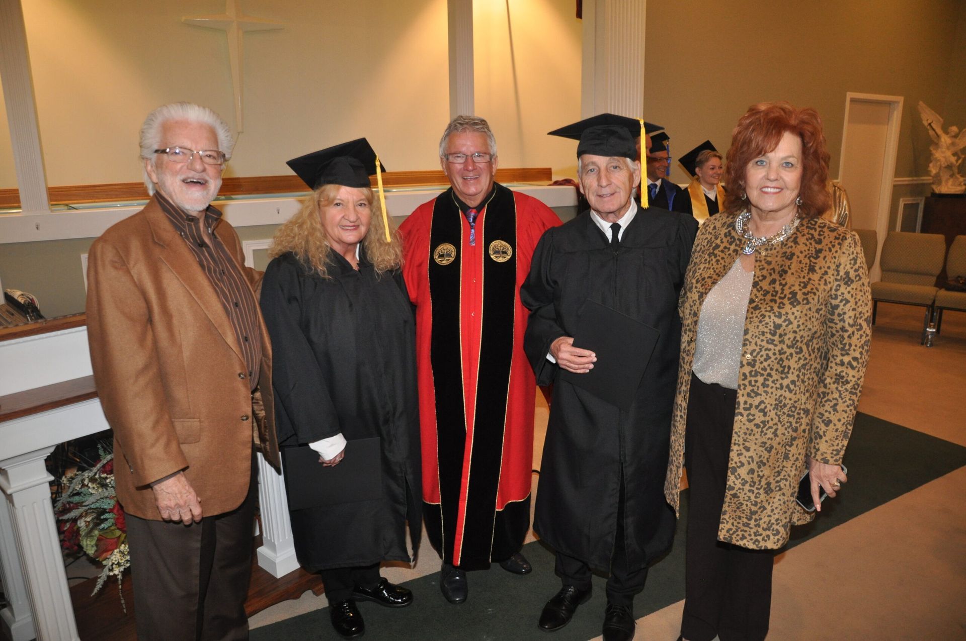 Five people in graduation attire, indoors. A robed man stands between two graduates and two others.