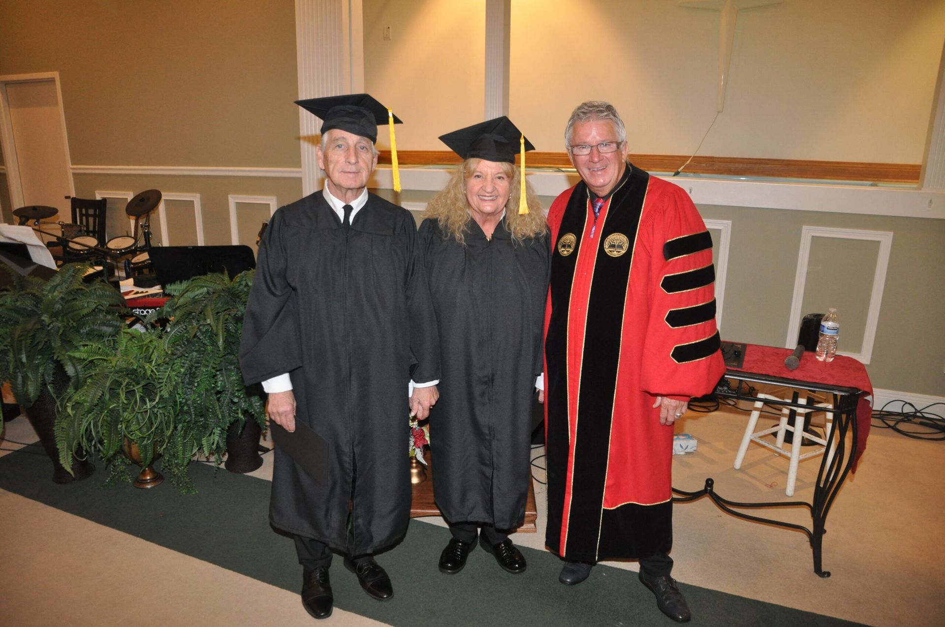 Three people in graduation attire pose indoors. Two in black gowns and mortarboards, one in a red robe.