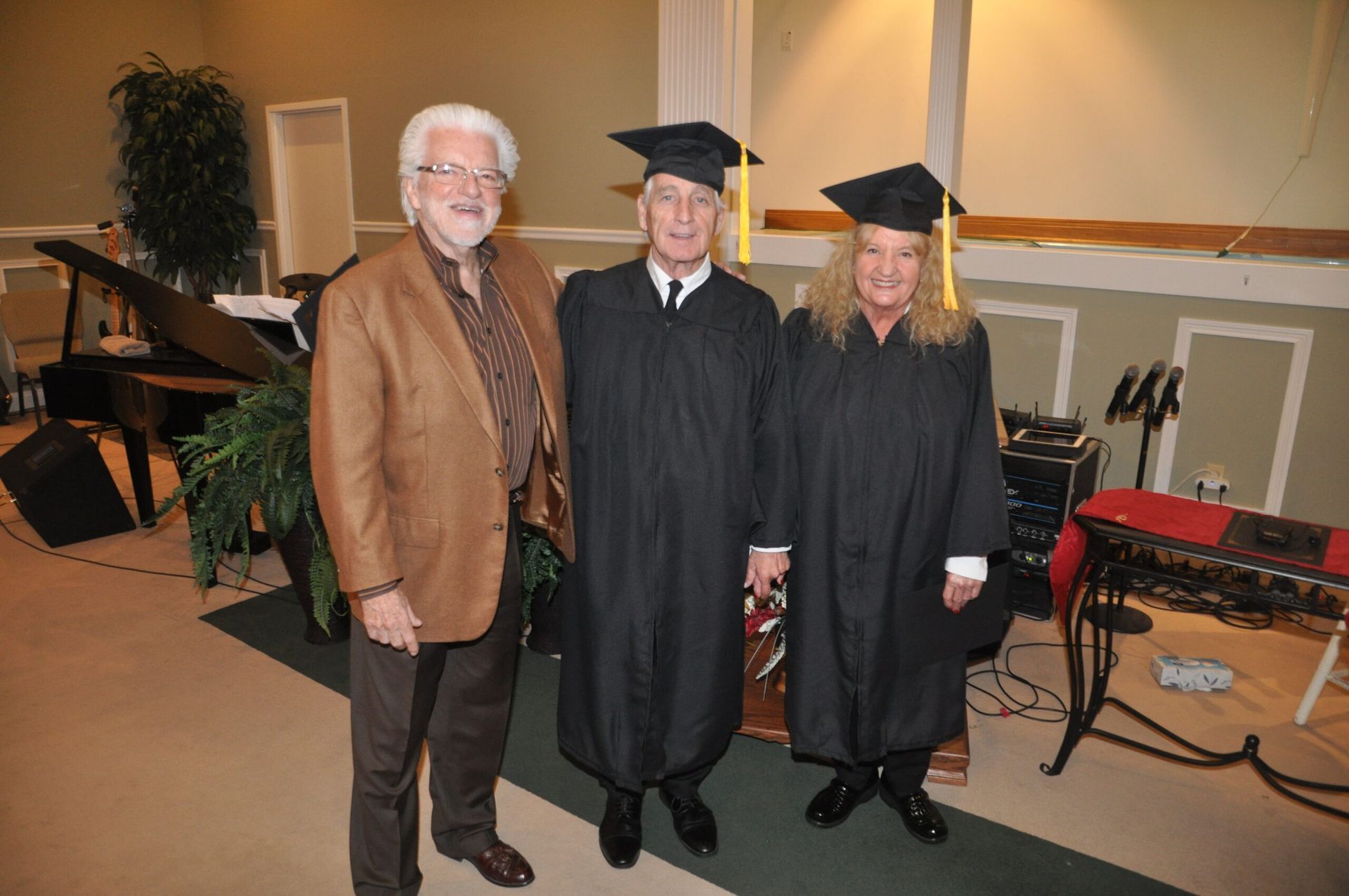 Three men pose indoors: one in a brown jacket, two in graduation gowns with caps.