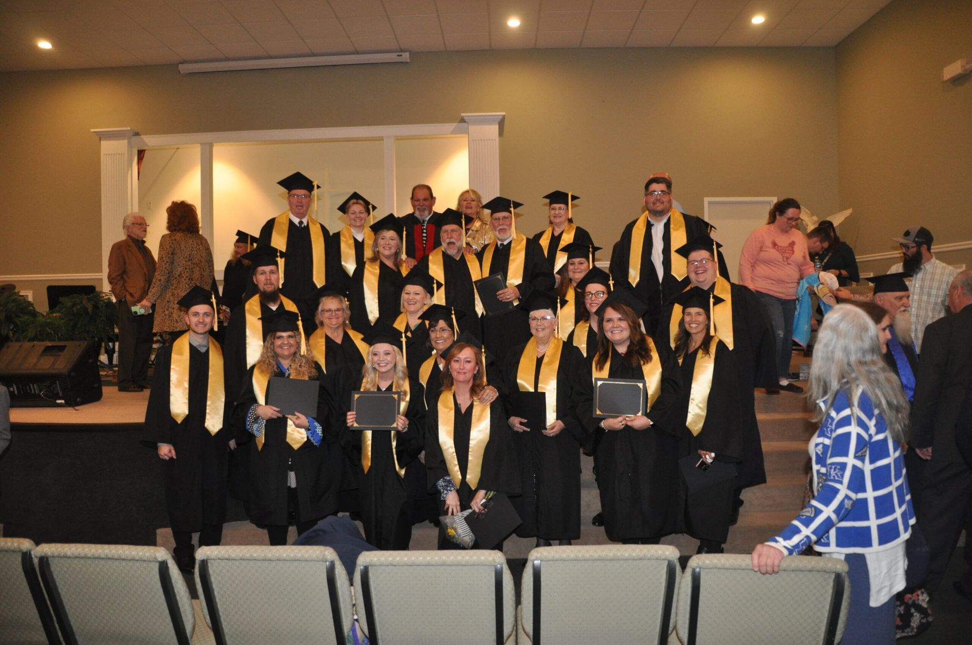 Graduates in black gowns and gold stoles pose on stage, holding diplomas.