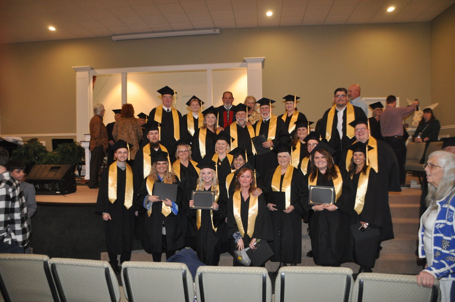 Graduation ceremony group photo, black gowns with gold stoles, holding diplomas, indoors.