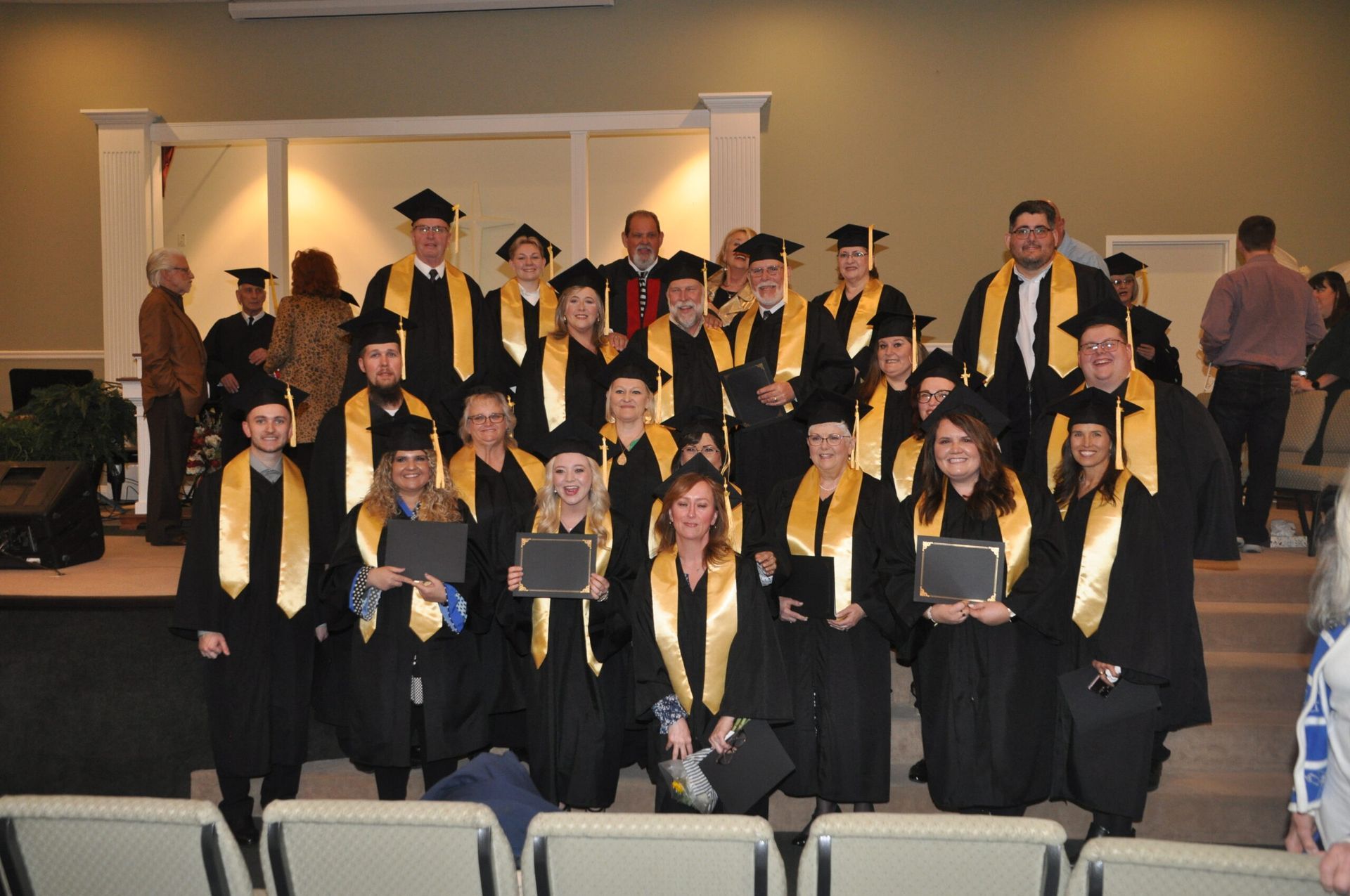 Group of graduates in black gowns with yellow stoles, posing on a stage.