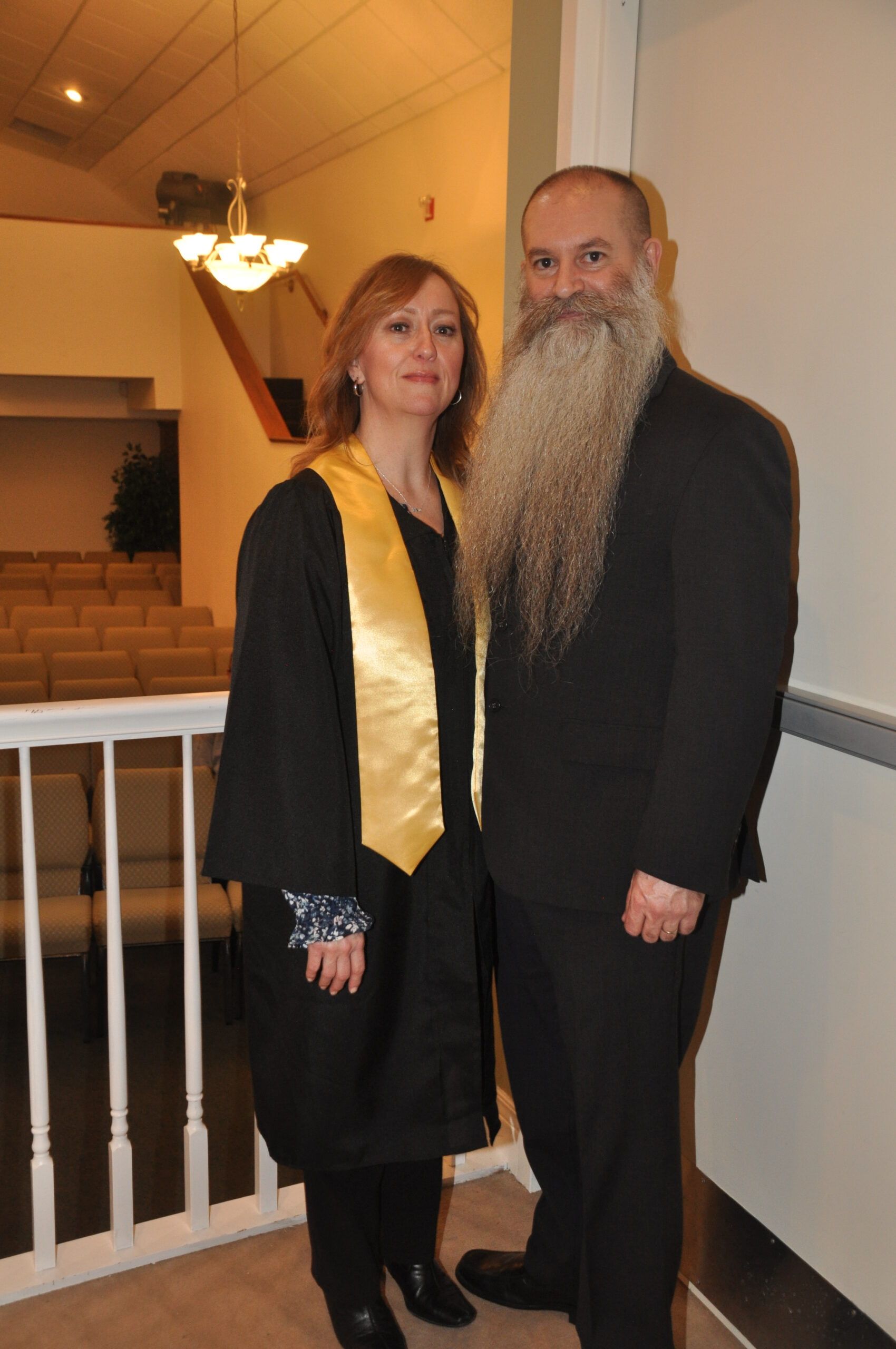 Woman in black robe with gold sash, man with long beard, pose near balcony in church.