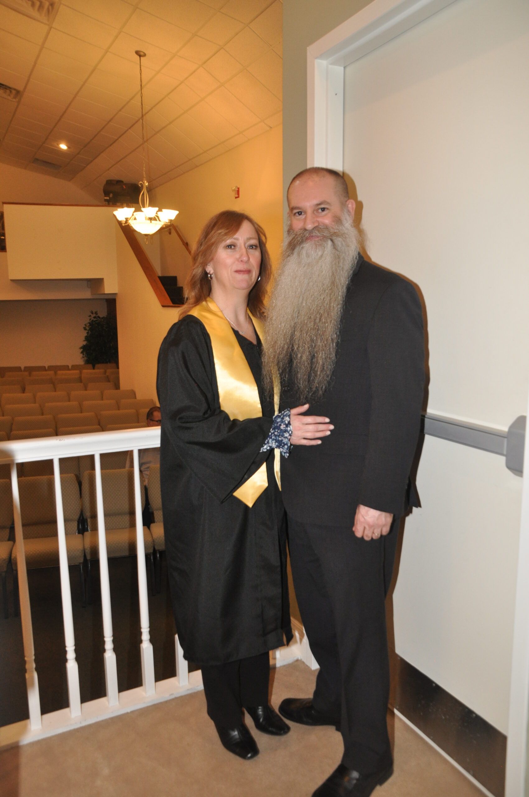 Woman in graduation gown and man with long beard pose indoors near a staircase.