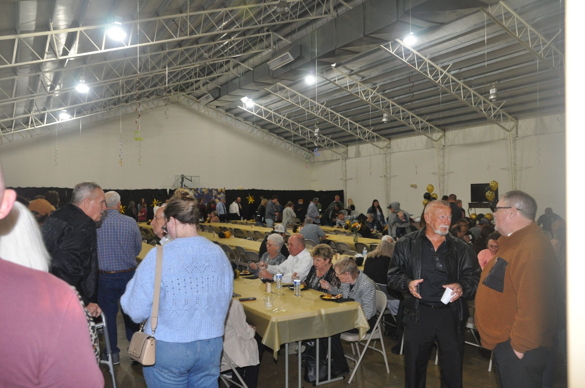 People gather inside a large hall at tables covered with gold tablecloths. Dim lighting.