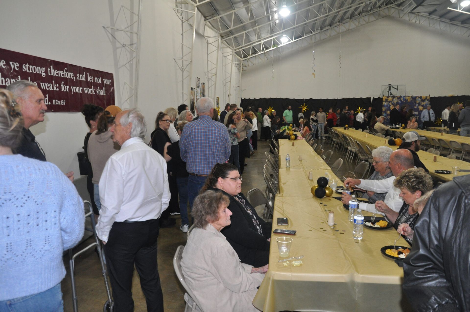 People gathered at long tables in a white, high-ceilinged hall. Some are eating, some are talking.