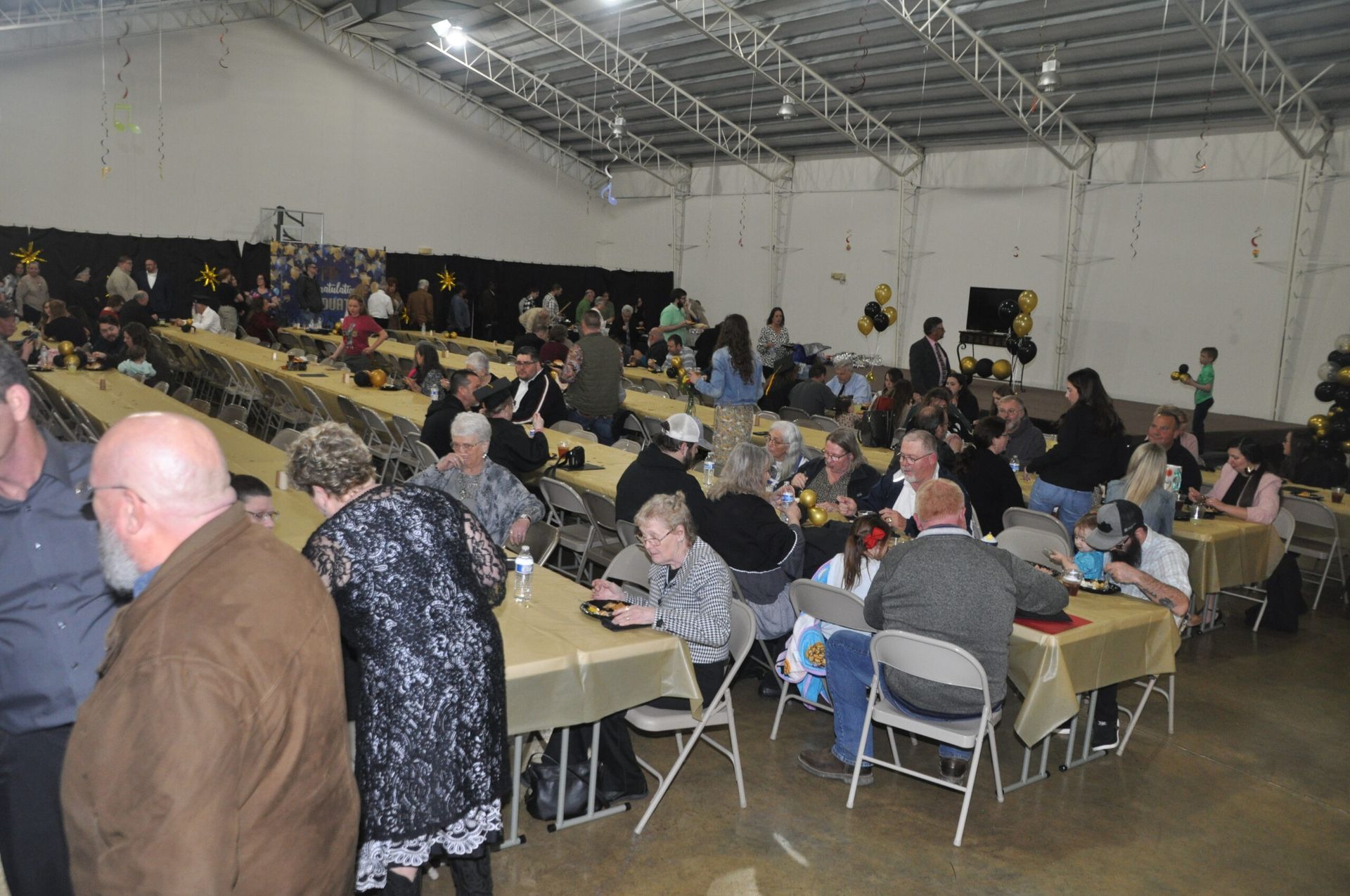 Large gathering in a hall with long tables covered in gold tablecloths; people socialize, eat, and move about.