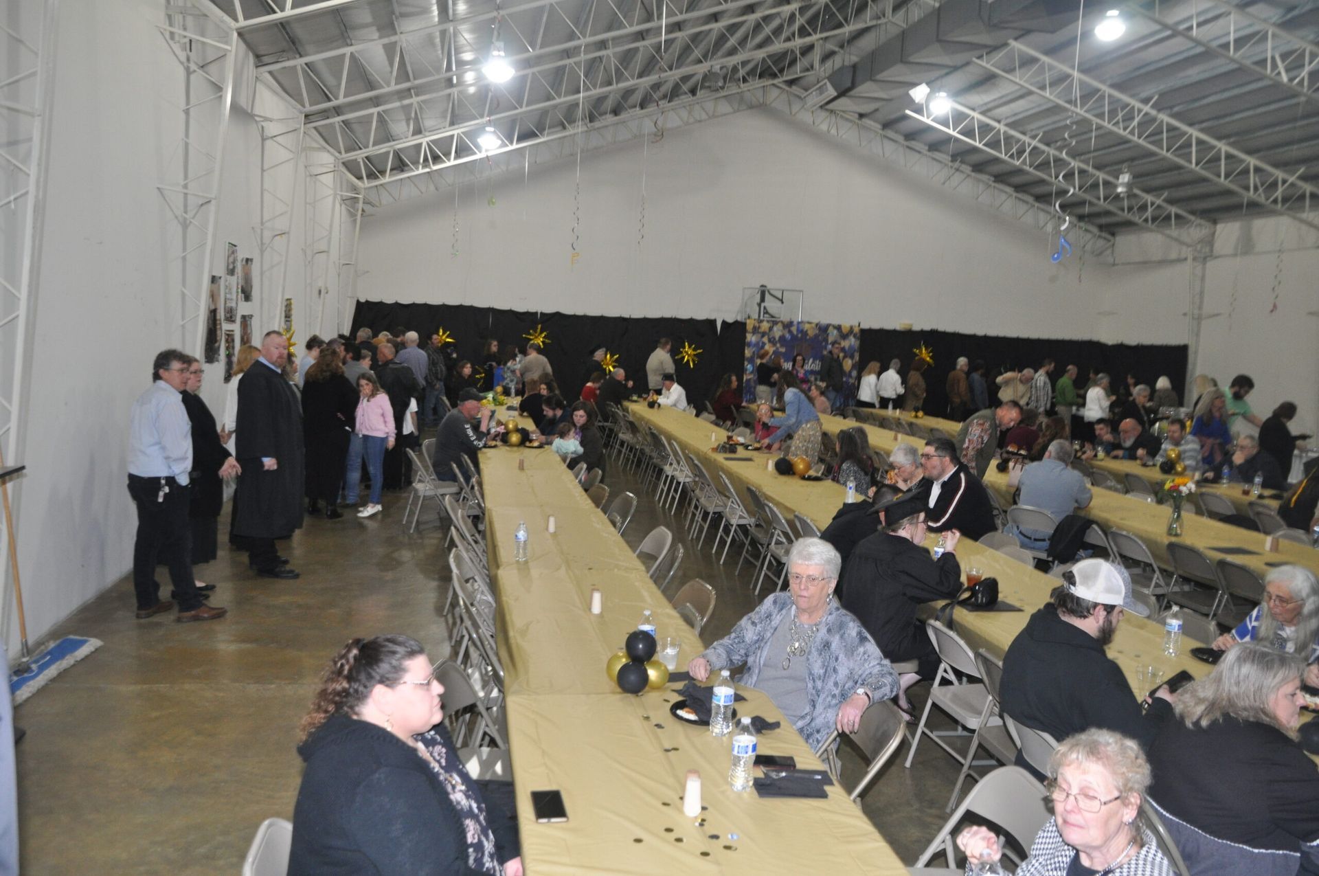 Large room with long tables set for a gathering. People sit at tables, others stand near the wall.