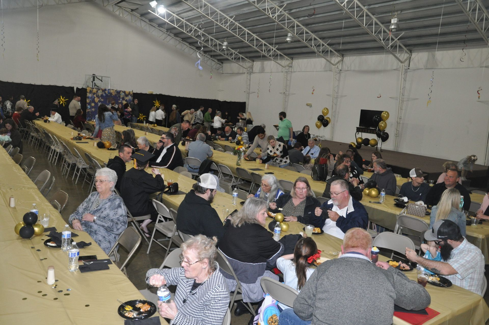 People seated at long tables in a large event space, some eating. Black and gold balloons decorate.