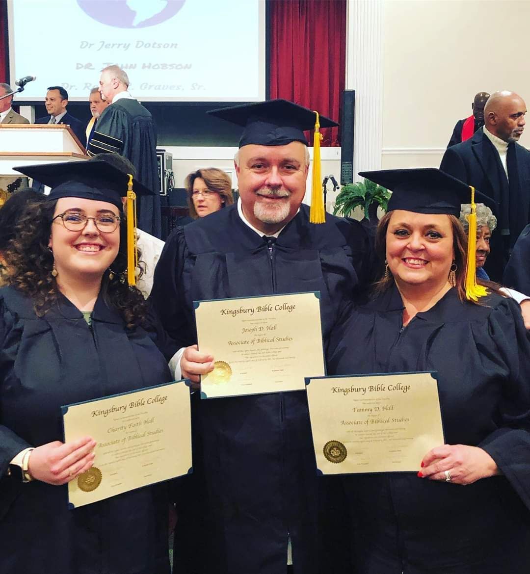 Three people in graduation gowns holding diplomas, smiling at the camera. Ceremony setting with a banner in the background.