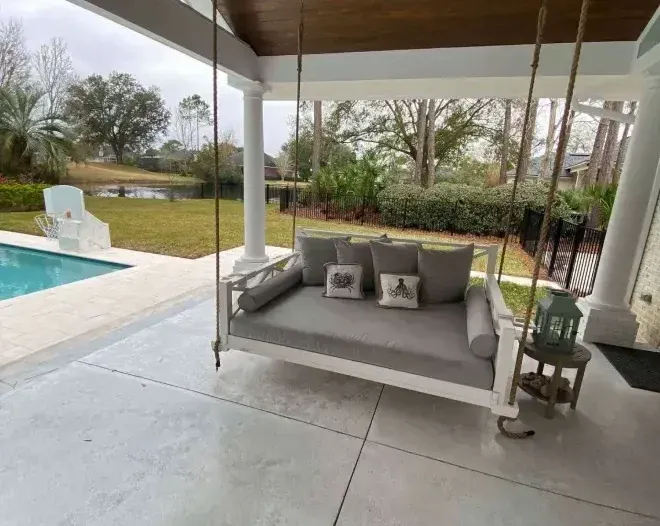 A white porch swing with gray cushions hangs on a patio overlooking a pool and yard.