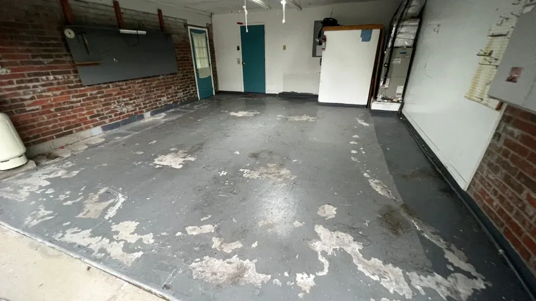 An empty, old garage with peeling gray paint on the floor, brick walls, and a teal door.