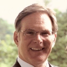 Man with glasses smiling, wearing a suit, outside with greenery in the background.