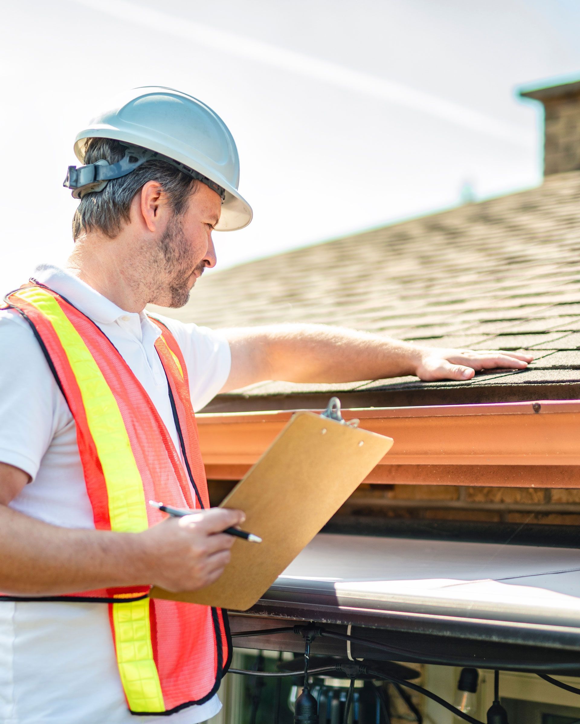 A man wearing a hard hat and safety vest is looking at a roof while holding a clipboard.