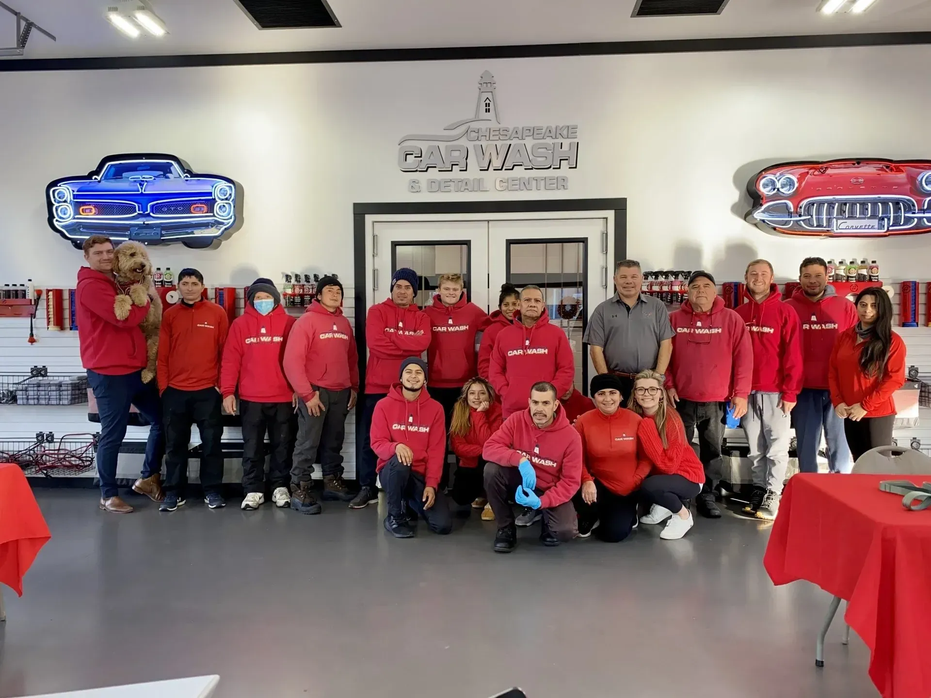 Group of people in red shirts posing inside a car showroom with red-covered tables and sports car wall signs.