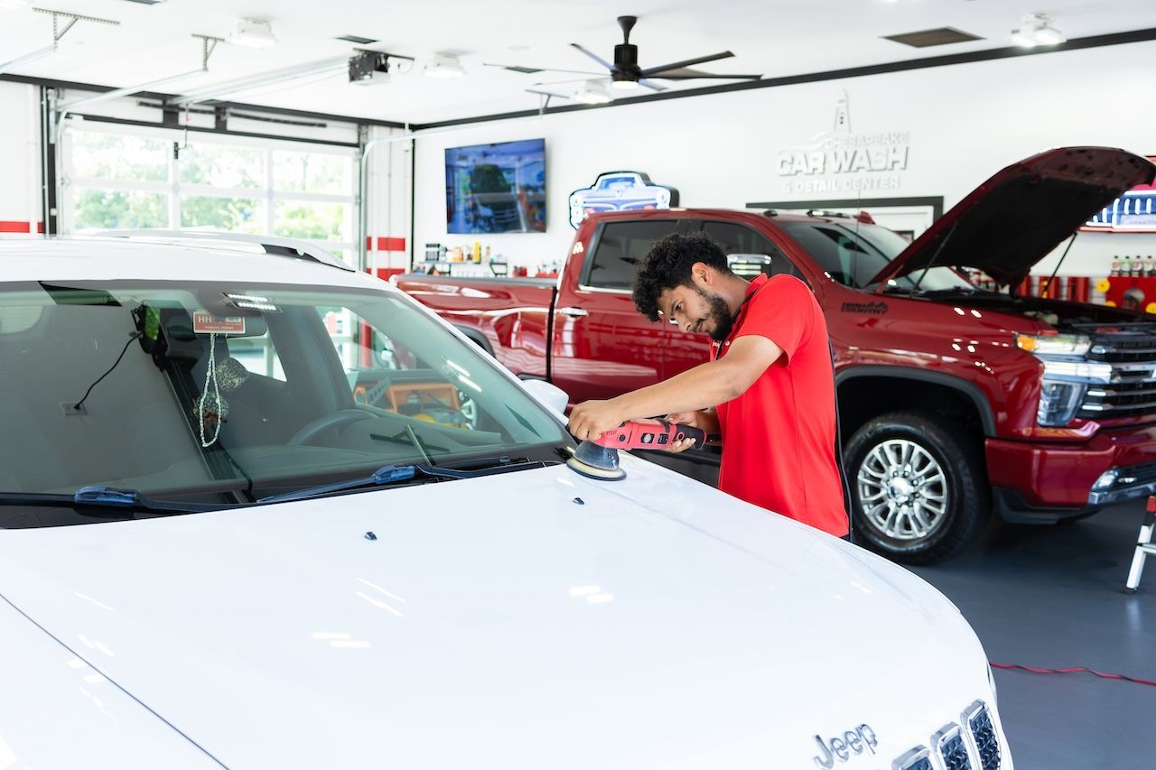 Person inspecting a white car in an auto repair shop, with a red SUV in the background.