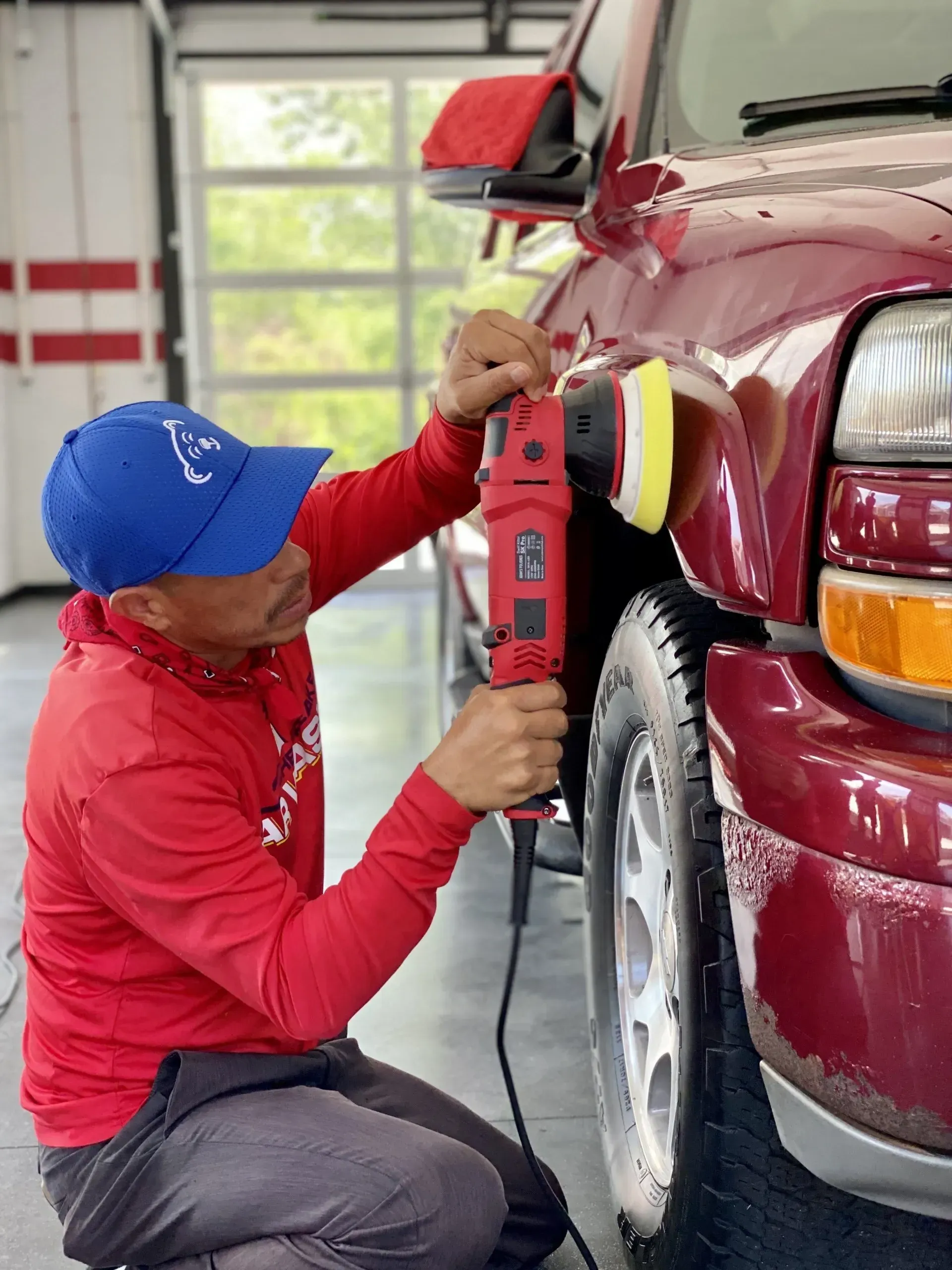 Person polishing a maroon SUV fender with a red buffer in a garage.