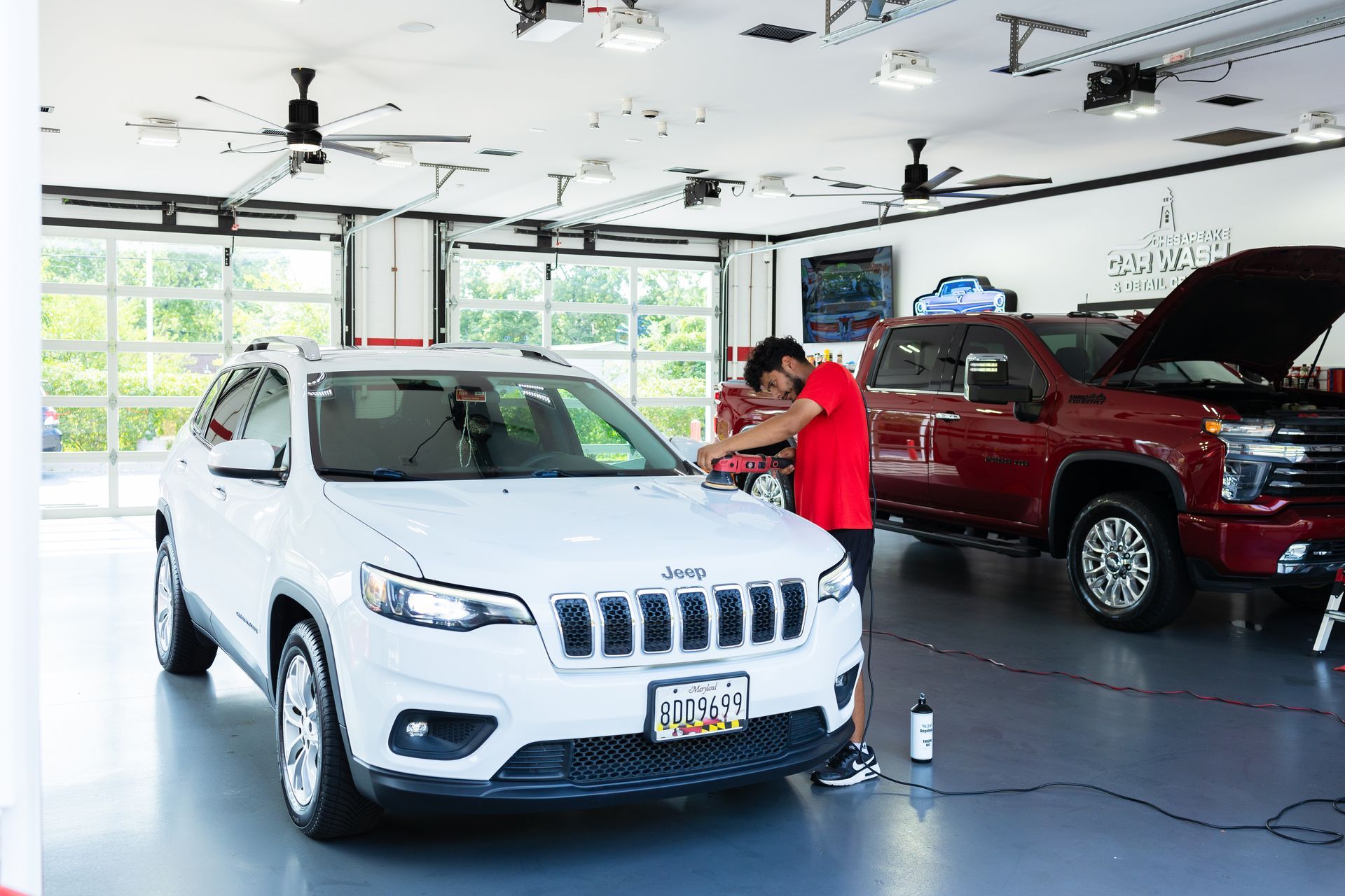 White SUV in a bright car showroom, with a person leaning on a red truck in the background.