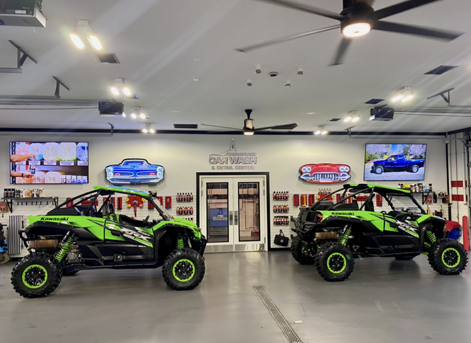 Two neon-green off-road buggies in a showroom with wall-mounted TVs and bright overhead lights