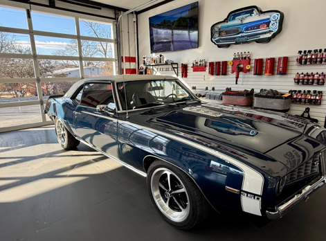 Classic dark blue muscle car in a bright garage with wall-mounted tools and a TV display.