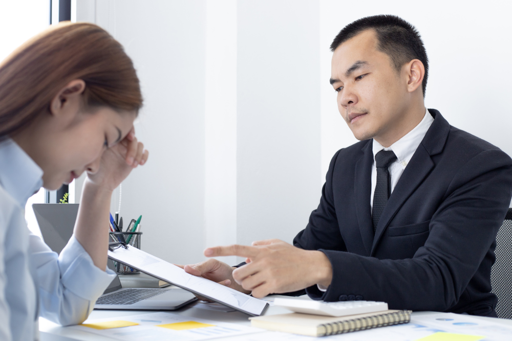 Man in suit pointing at document, woman with head in hand, at desk.
