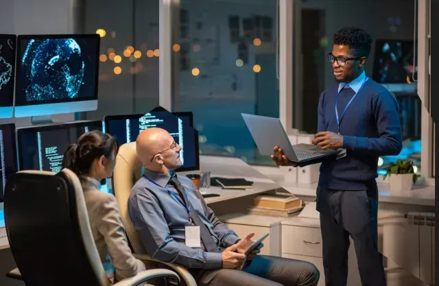 A Black man presents with a laptop to colleagues in a tech office at night.