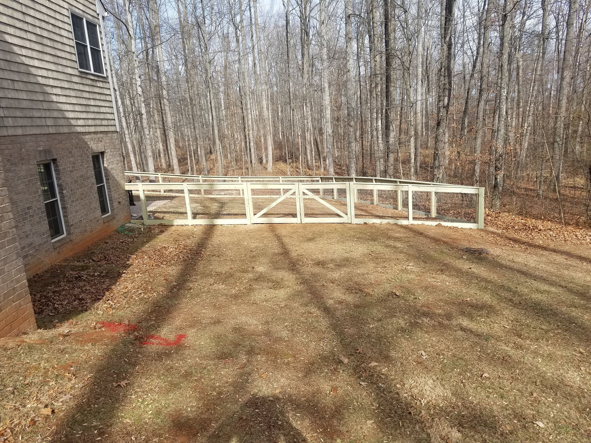 A partially built wooden fence extends from a brick house into a wooded area on a sunny day.