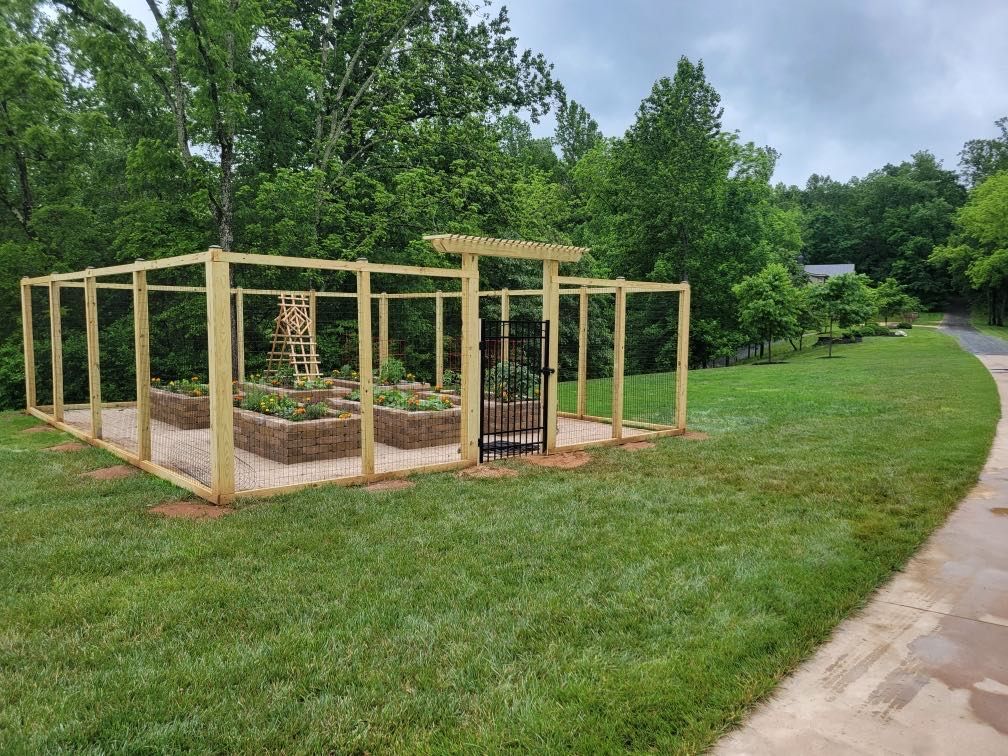 Raised garden beds within a wooden fence, with a gated entrance and a pergola, in a grassy area.