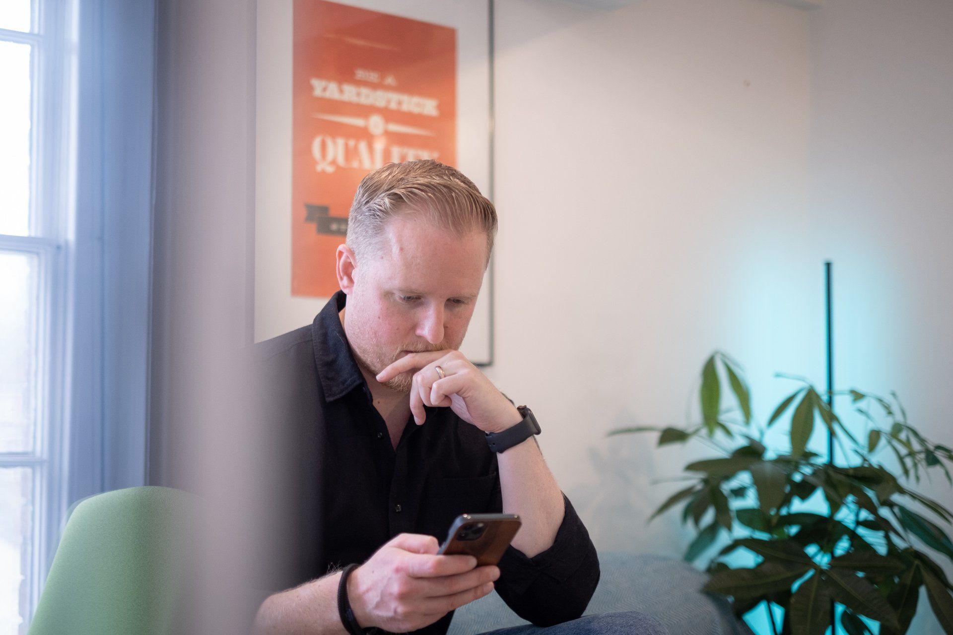 Russ, a white male with light hair wearing a black shirt and watch sits at a desk, looking at a phone in his hand.