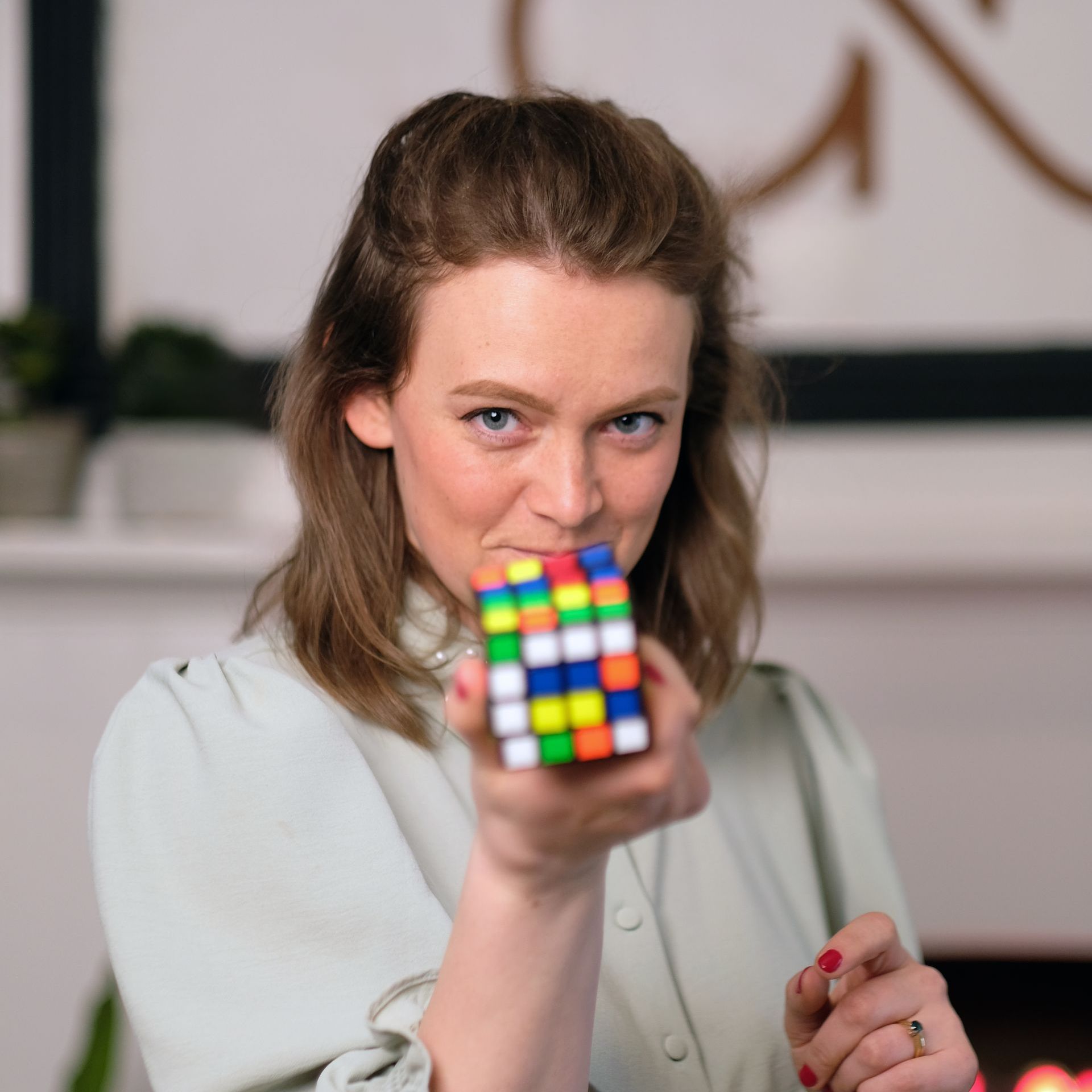Headshot: a white woman with light brown hair holds up a rubik's cube to the camera wearing a blue-grey top with embellished collar