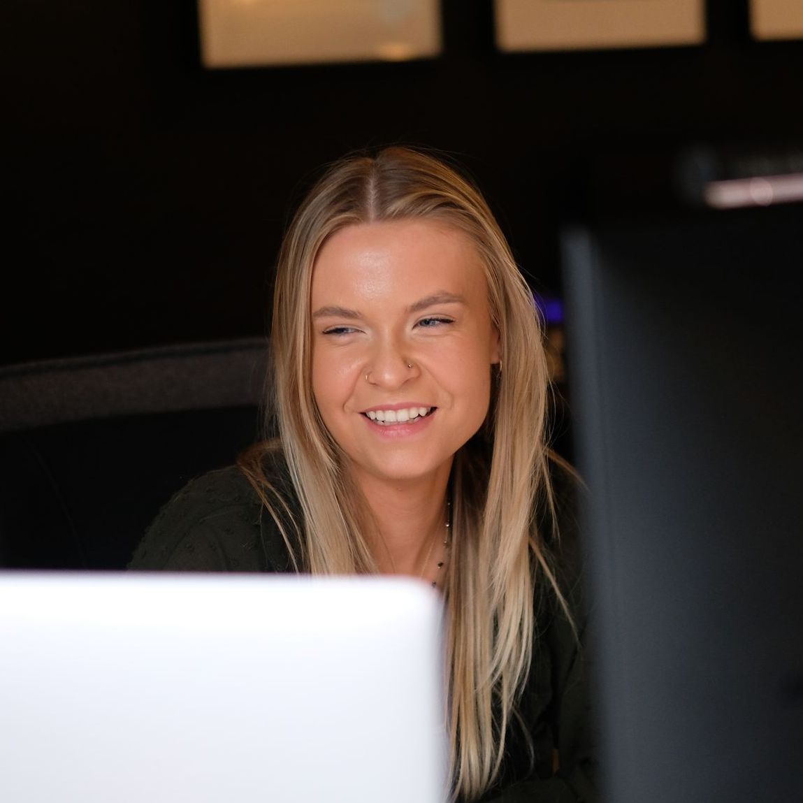 Headshot:  a young white woman with blond hair smiles into the corner of the room, sat behind a blurred out computer screen in the foreground.