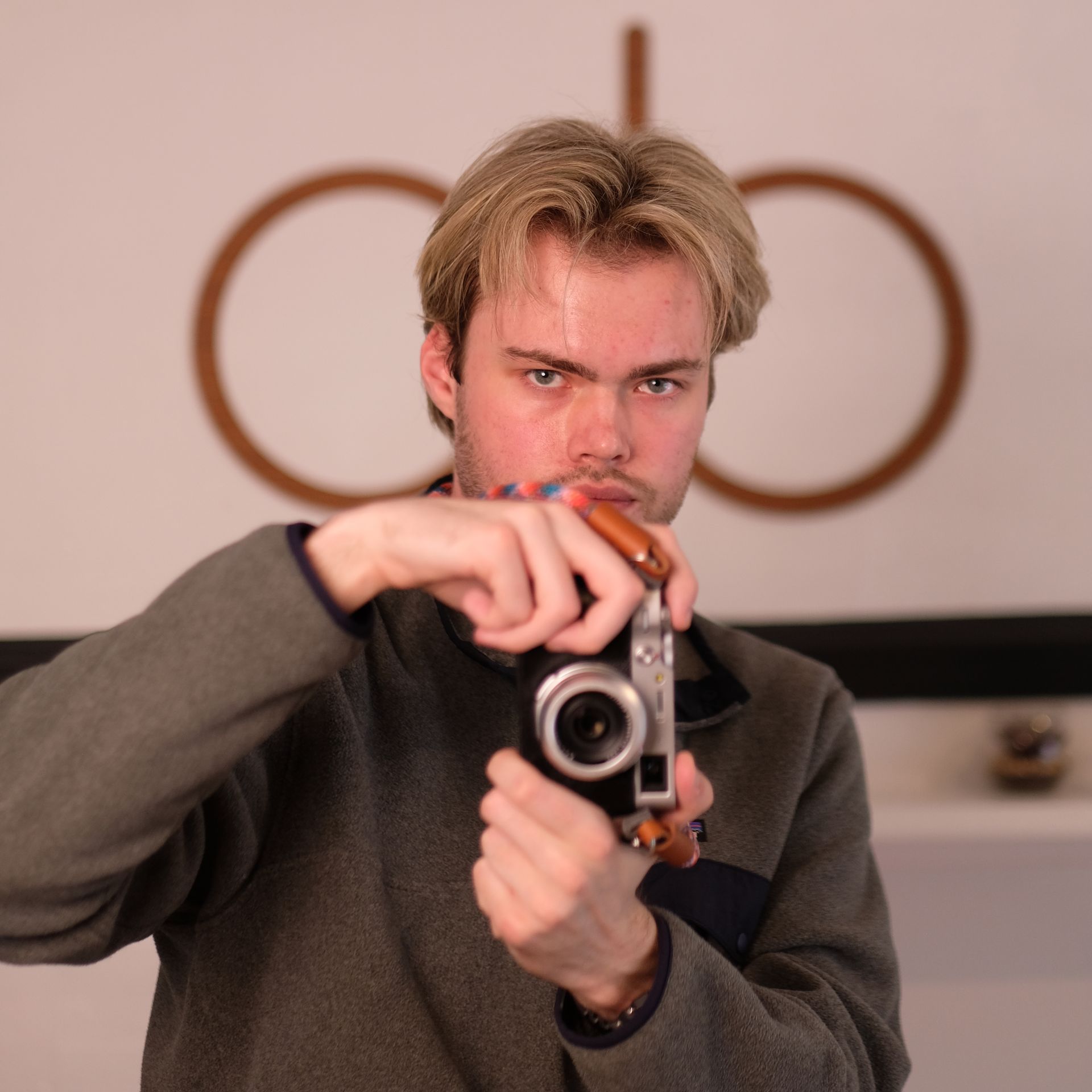 Headshot: a white man with blond hair and stubble holds up a retro camera and looks into the shot, wearing a fleece with a zip