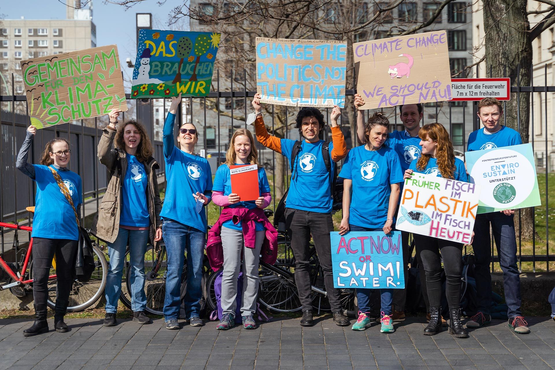 A varied group of people, all wearing blue t-shirts with the earth in white on them stand in front of a tall metal fence which has some biked chained to it. They hold a variety of homemade posters demanding climate action in both English and German.