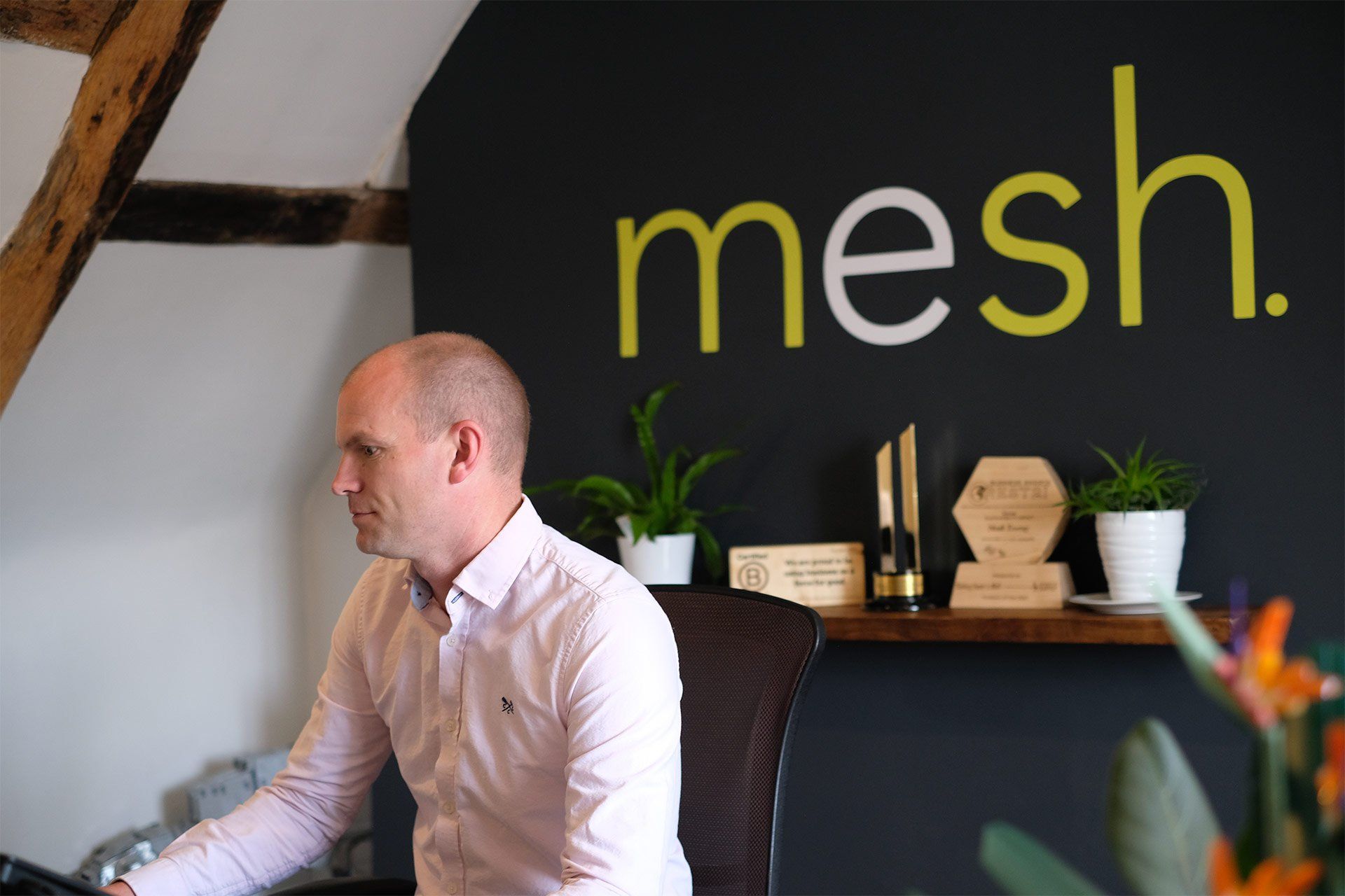 Mesh Energy director Doug, a white man, sits at a desk , concentrating on the screen in front of him. The mesh logo is painted on the wall behind him and several awards are visible on a shelf, including a B-corp award.