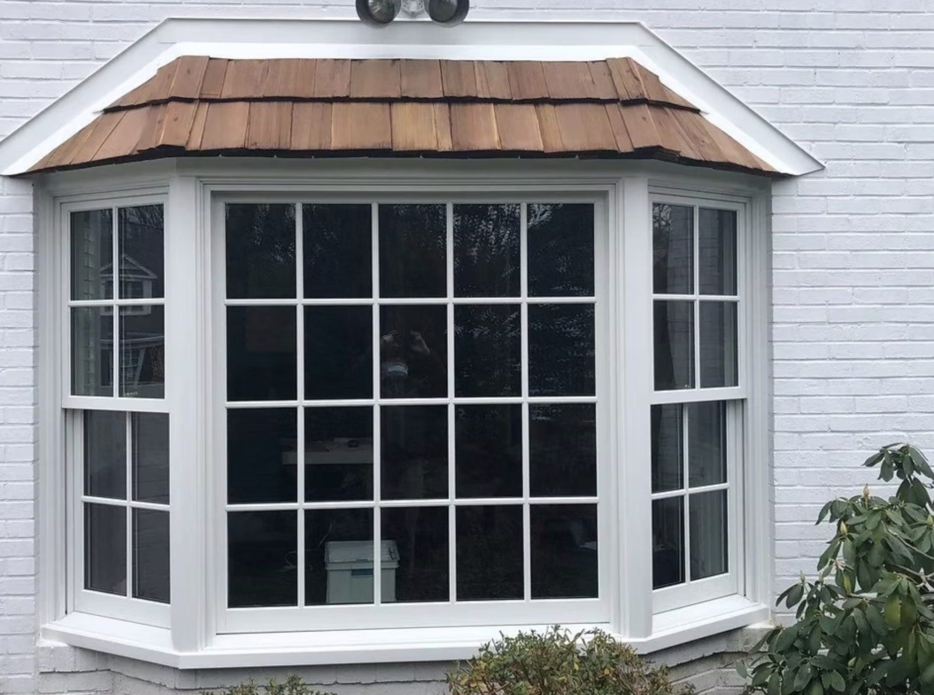 A white bay window with a wooden roof on a white brick house.