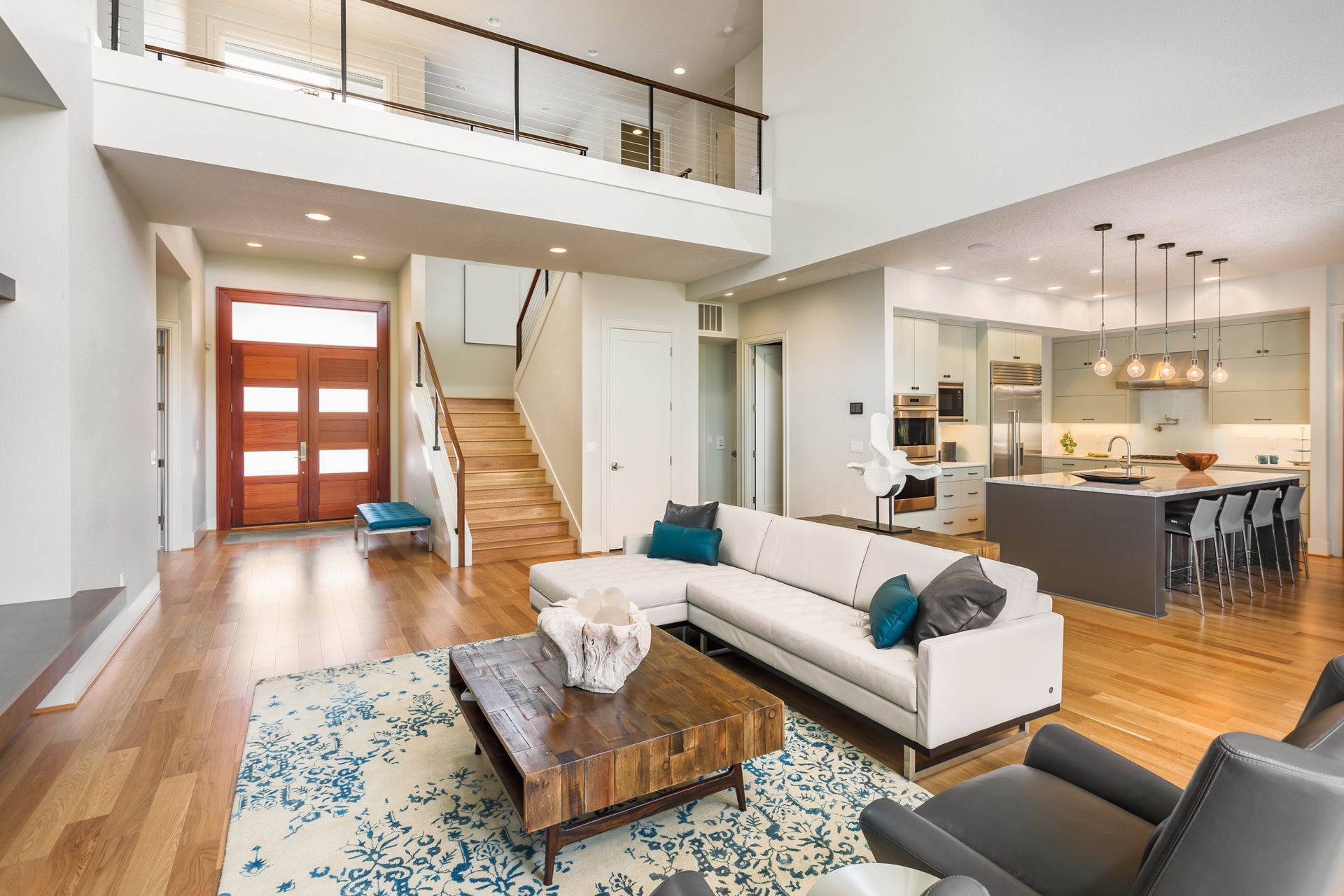 Modern living room with a kitchen and staircase, featuring a white sofa, wooden floor, and a blue rug.