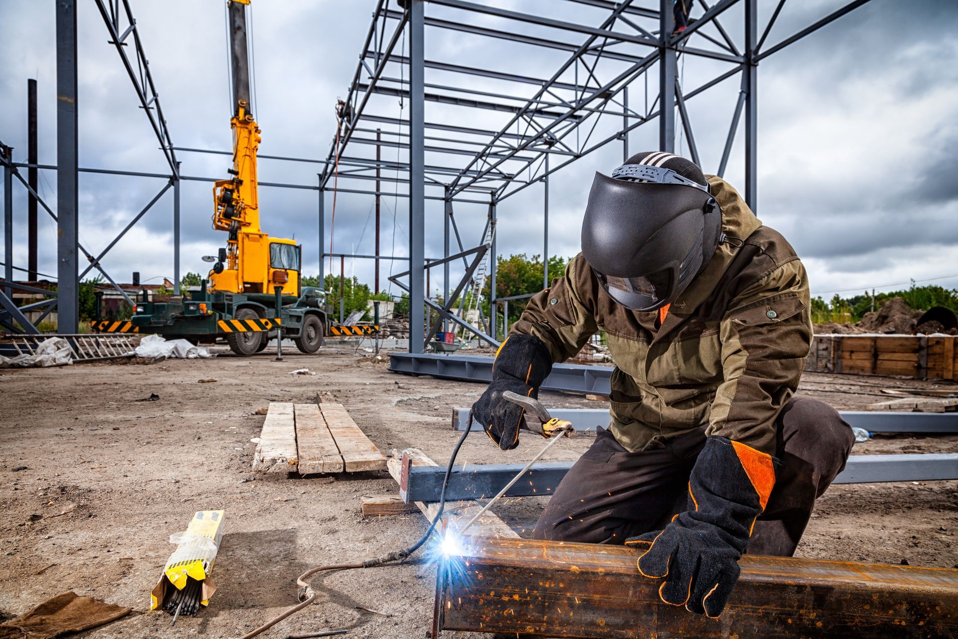 Welder wearing a helmet working on steel beams at a construction site with a crane in the background.