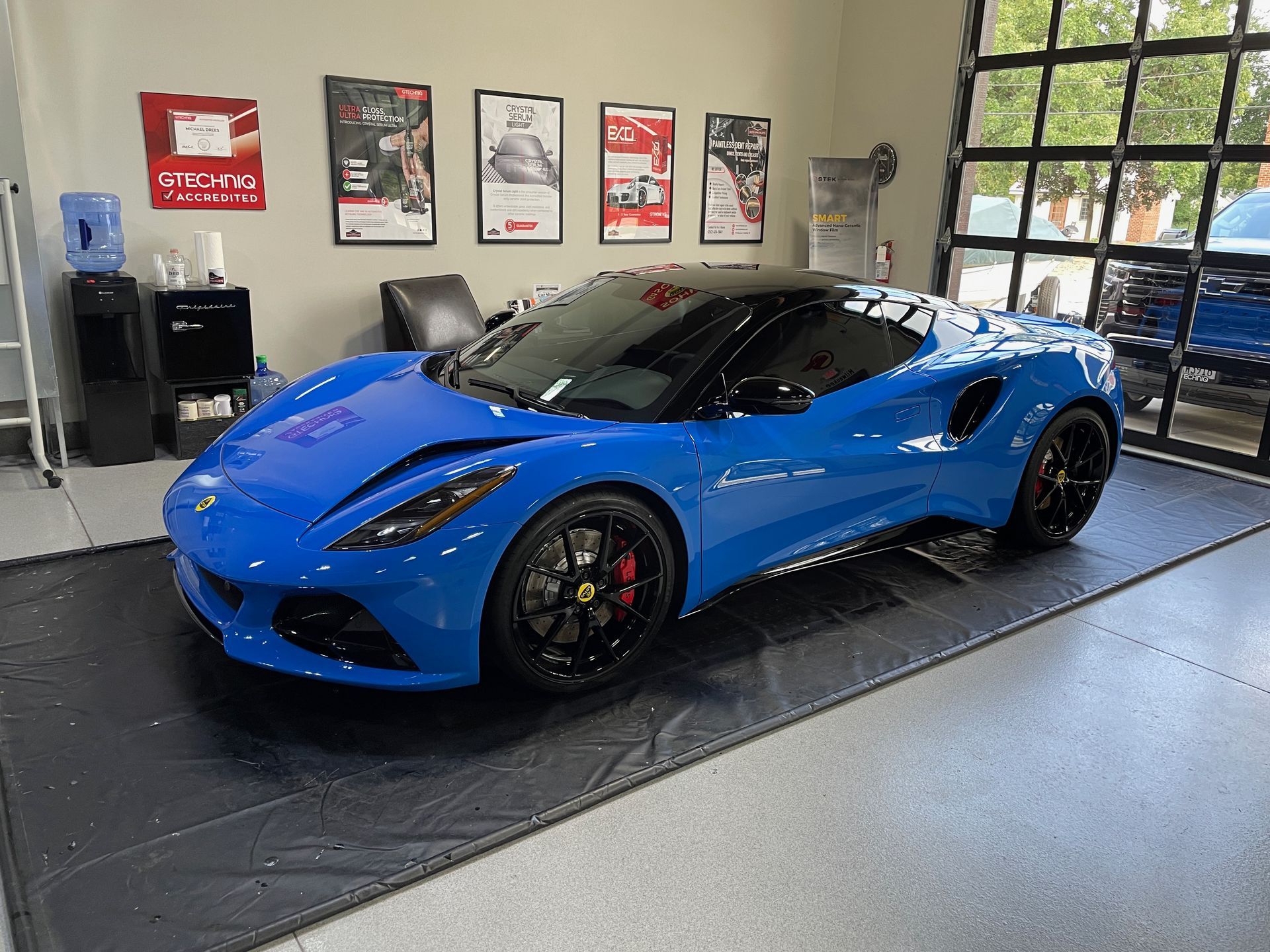 Blue sports car on a black mat inside a shop with posters on the wall and a glass garage door.