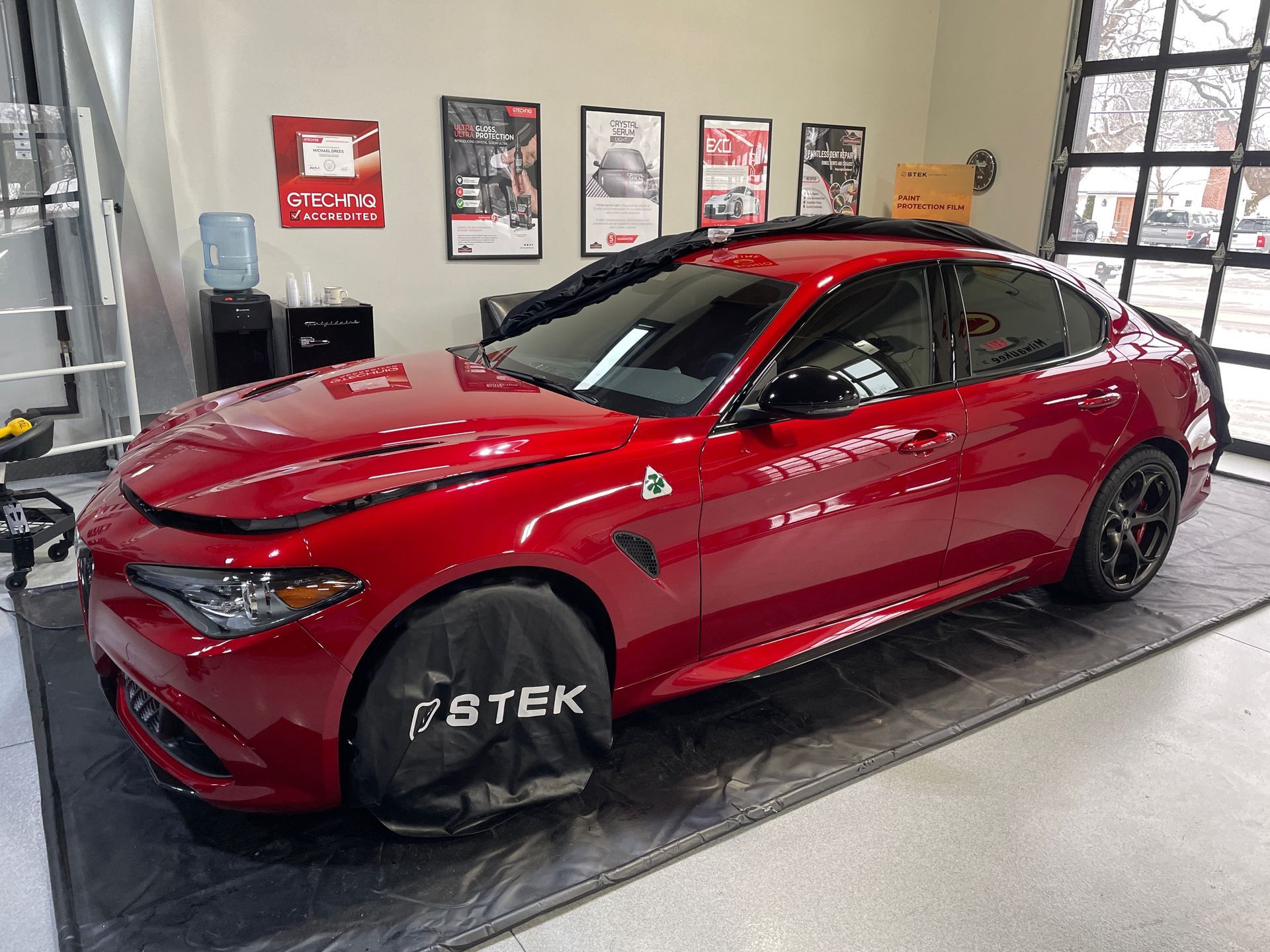 Red Alfa Romeo sports car in a garage with wheel cover, near posters and a window.