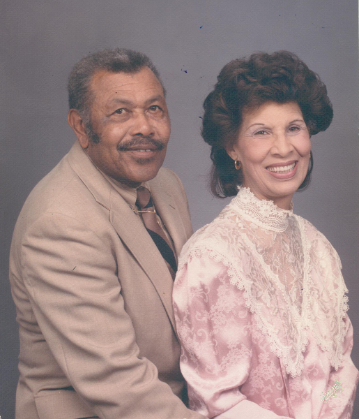 Older Black couple, man in tan suit, woman in pink lace dress, smiling, studio portrait.