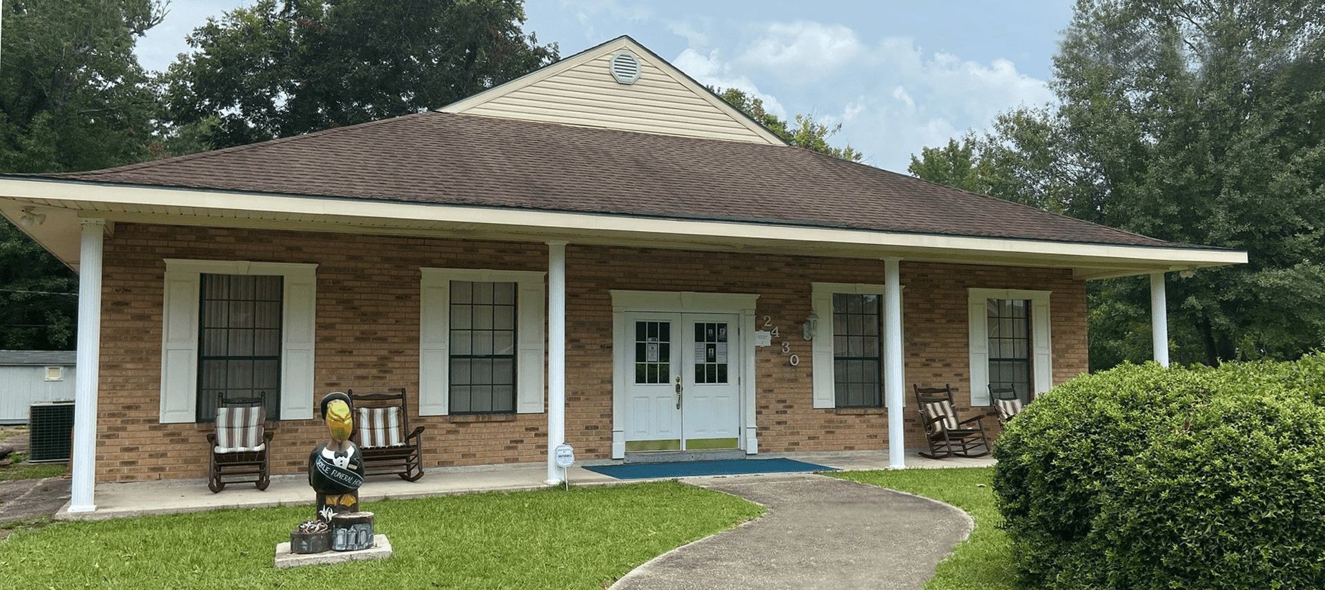 Brick building with white pillars, windows, and a brown roof. Two rocking chairs and a statue on the porch.