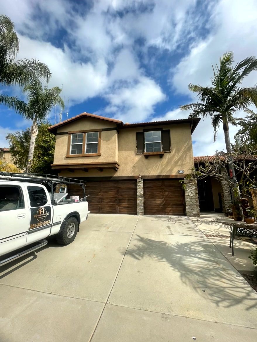 Two-story house with brown garage doors, tan stucco, and palm trees. A white truck is parked in the driveway.