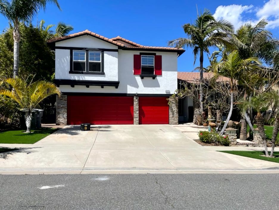 Two-story house with red garage doors and shutters. Palm trees frame the white facade and driveway.