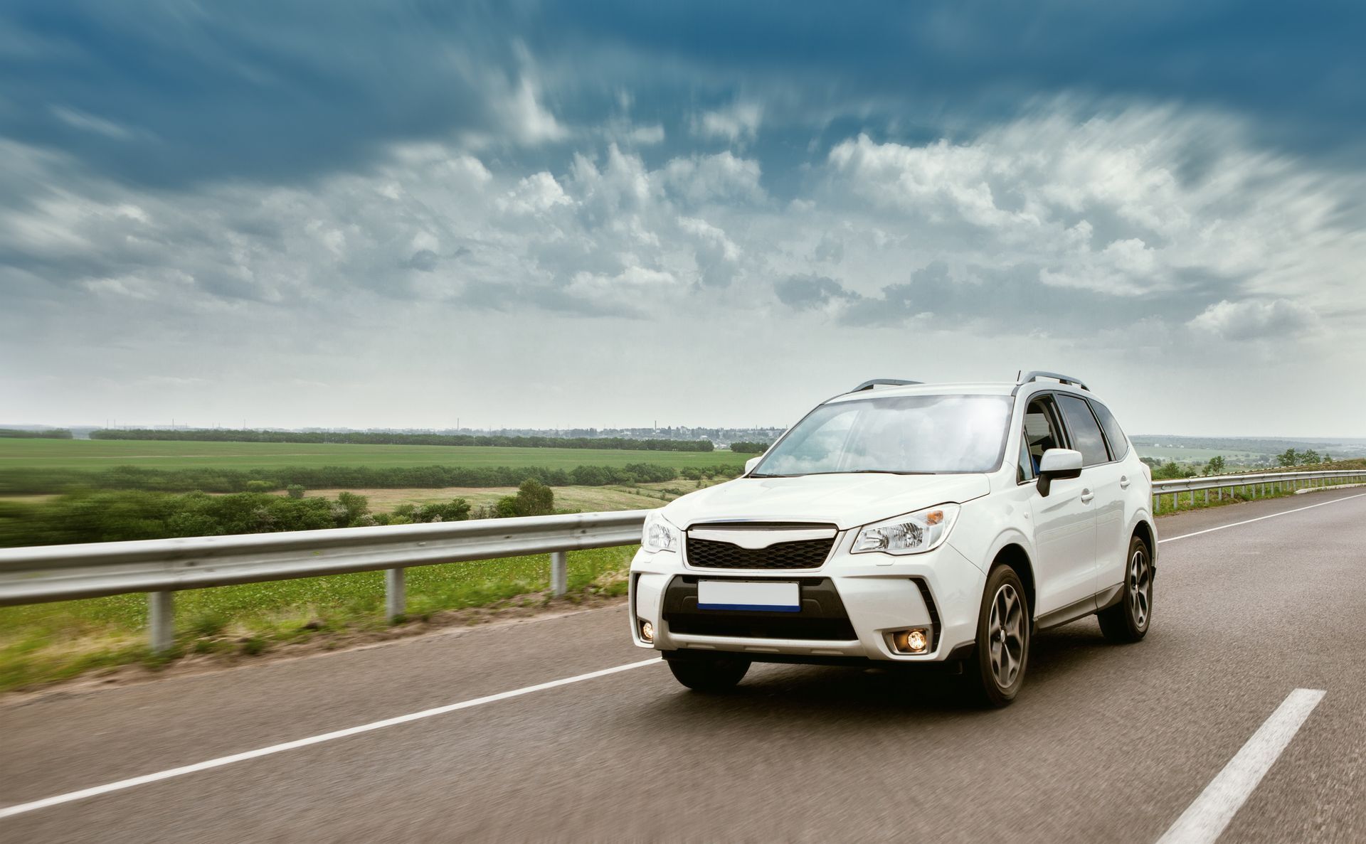 White SUV driving on a paved road, countryside background, cloudy sky.