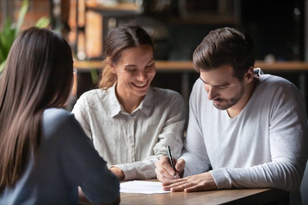Couple signing documents at a table, smiling. Woman in light shirt. Another woman faces them. Indoors, warm light.