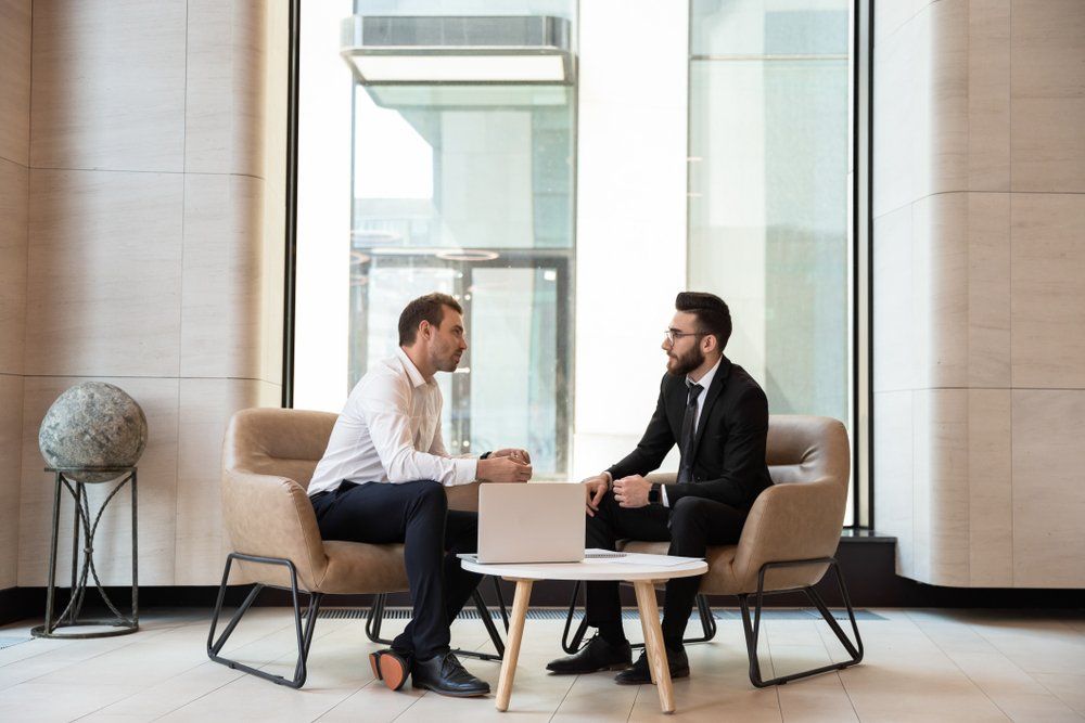 Two men are sitting at a table with a laptop and talking to each other.